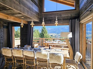 Un intérieur en bois avec une table à manger dressée pour quatre personnes, des chaises en osier agrémentées de plaids en fourrure, et une vue sur les montagnes par de grandes fenêtres.