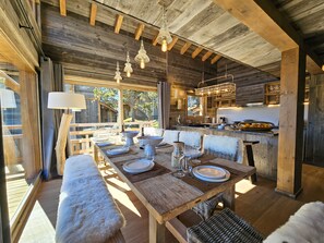 Un intérieur rustique en bois, avec un coin repas, des poutres apparentes et une vue sur l’extérieur.