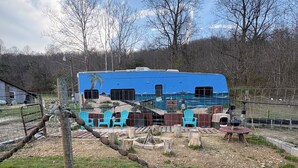 A blue RV with a palm tree mural, surrounded by a fenced area with chairs and a fire pit.