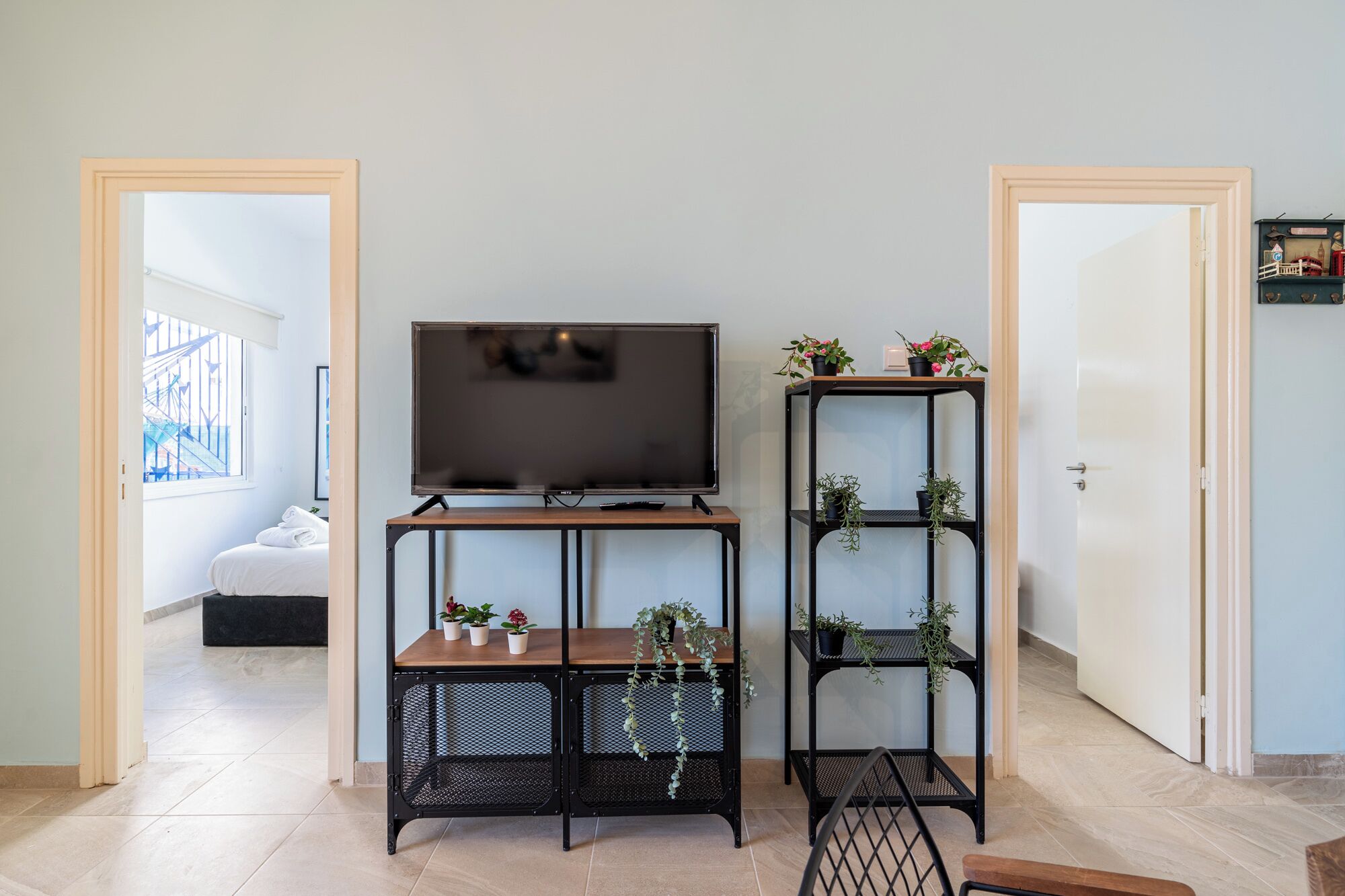 A modern living room with a TV on a wooden stand, two black metal shelves with plants, and a view of a balcony through a window.