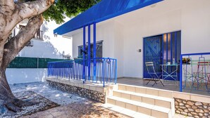 A covered outdoor seating area with a stone-tiled floor, a blue railing, and a staircase leading to a door with a geometric design. - Nala - 2 Bedroom Apt & Patio in Larnaca (Larnaca)