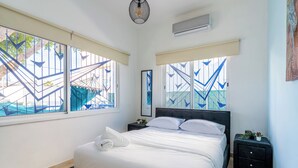 A neatly made bed with white linens, a bedside table with a plant, and a window with geometric patterns. - Nala - 2 Bedroom Apt & Patio in Larnaca (Larnaca)