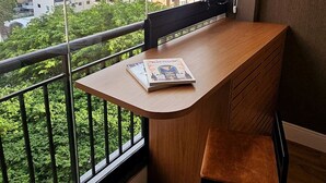 Een modern appartement met een houten bureau en stoel, grote ramen met uitzicht op stadsgebouwen, en een balkon met beplanting.