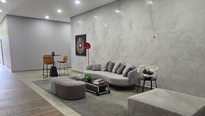 Een moderne woonkamer met een grijze bank, twee fauteuils, een salontafel en een bijzettafeltje. Er hangt een schilderij aan de muur en staat een vloerlamp.