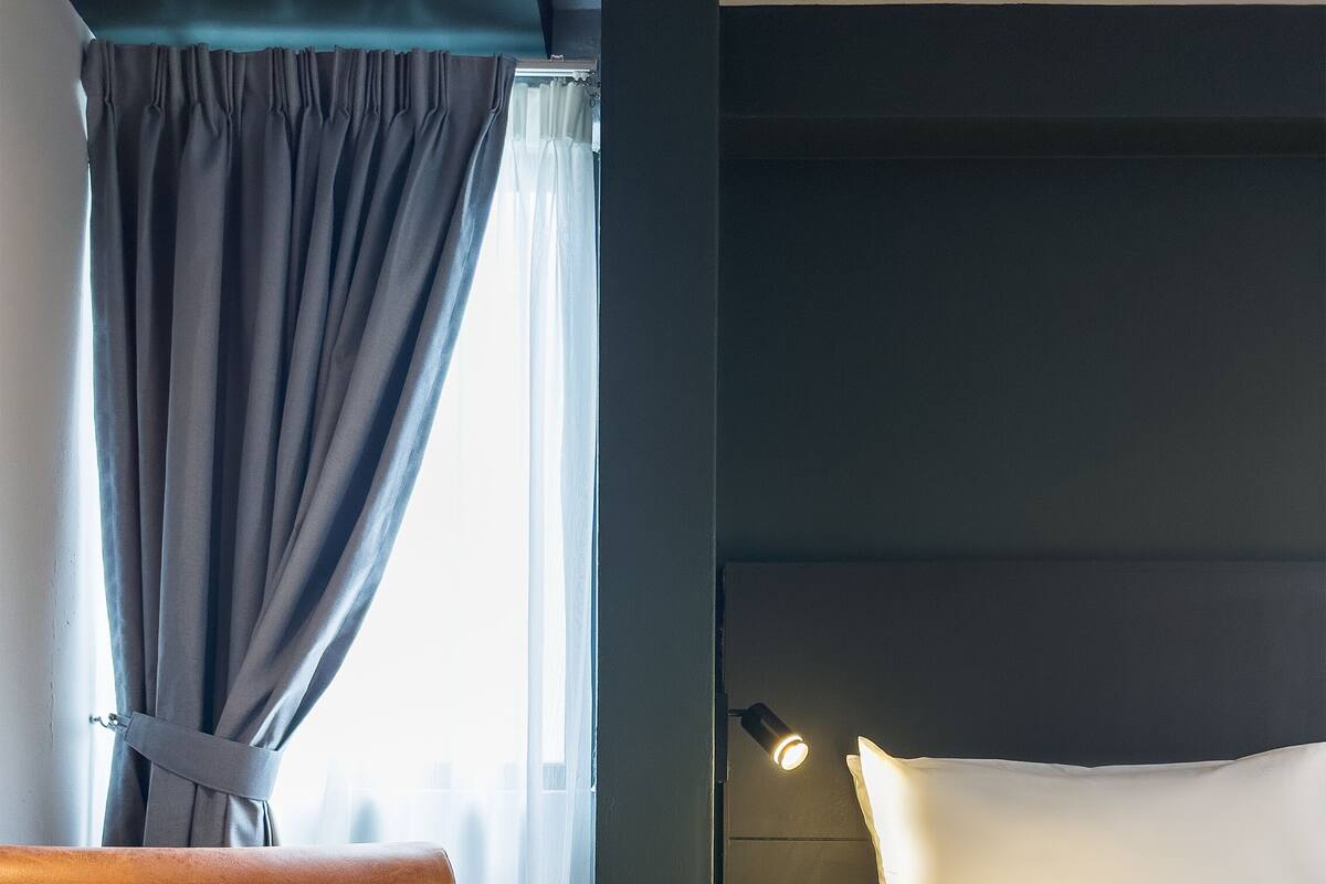 A hotel room with a brown armchair, a bed with white linens, a window with curtains, and a wall-mounted lamp.