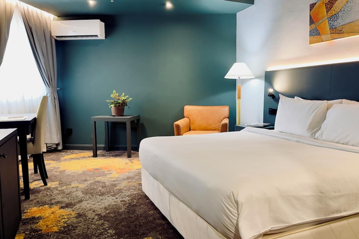 A hotel room with a large bed, a desk, a chair, a small table with a plant, and a wall-mounted lamp.