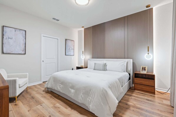 A modern bedroom with a large bed, a white chair, wooden flooring, and two bedside tables with lamps. - Epic Family Villa Pool Theater Themed Bedrooms (Clermont)