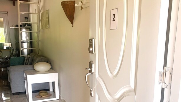 Une chambre d’hôtel avec un sol en marbre, une porte blanche portant un numéro, une petite table blanche et un canapé avec des coussins.