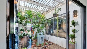 A glass-enclosed patio with various plants, a seating area with cushions, and a stone pathway. - Luxury Retreat ✨ Views, Heated Pool+ Spa Baths.♥️smart bidet (Palm Desert)