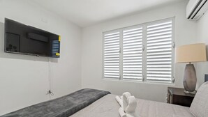A hotel room with a bed, a TV mounted on the wall, a window with blinds, a bedside lamp, and a small table. - Comfortable Home Prime PR Location 2-br Condo (Ceiba)