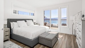 Ein Schlafzimmer mit einem großen Bett, einem Nachttisch, einer Kommode und Blick auf einen See und Berge.
