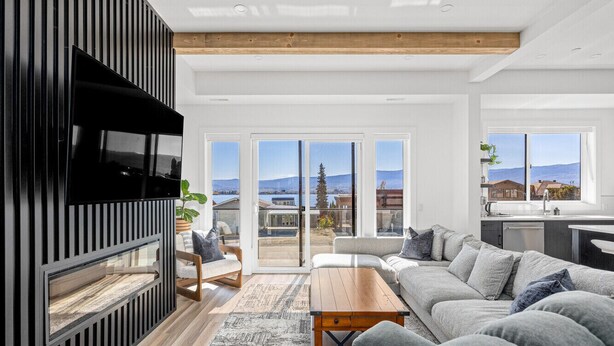 Ein modernes Wohnzimmer mit einem Ecksofa, einem Holztisch und einem Kamin. Große Fenster bieten einen Blick ins Freie.