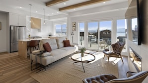 A spacious living area with a sofa, coffee table, and chairs, a kitchen with a refrigerator and pendant lights, and large windows offering a view of the outdoors.