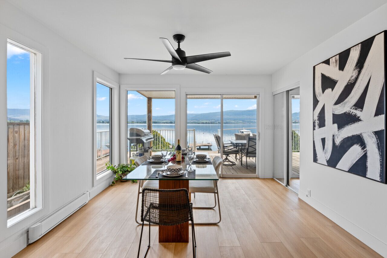 Un comedor amplio con una mesa de vidrio, sillas y un ventilador de techo. Ventanas grandes ofrecen vistas al exterior.