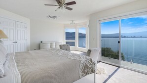 Un dormitorio con cama, cómoda, silla, ventilador de techo y una ventana grande con vista a un lago y montañas.