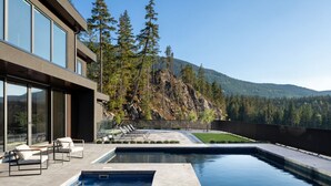 Una casa moderna con piscina, mobiliario exterior y vistas a una montaña boscosa.