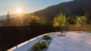 Un espacio exterior empedrado con una cerca metálica curva, pequeños árboles y montañas al fondo durante el atardecer.