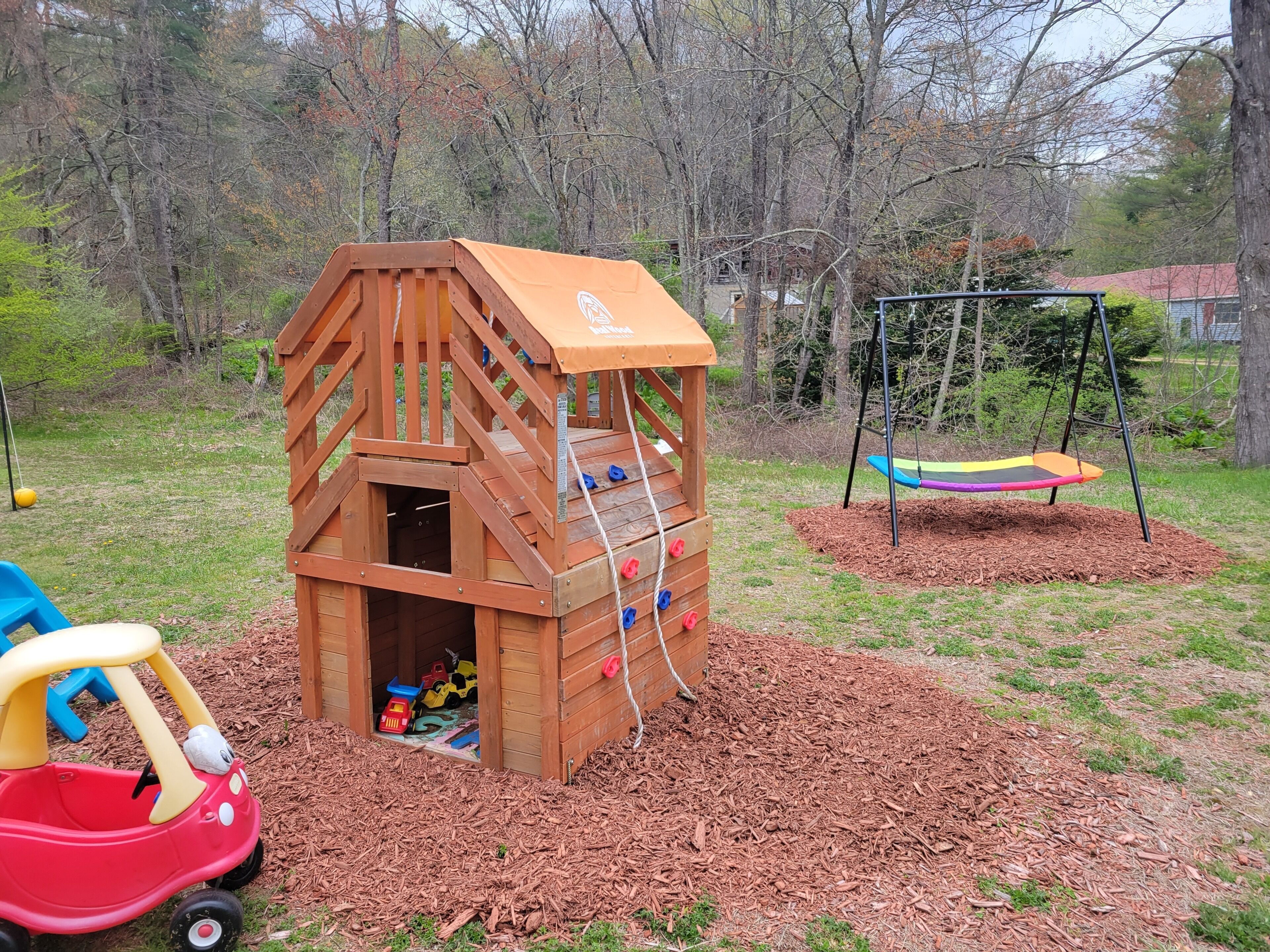 A wooden playhouse with a climbing wall, a swing set, and a toy car.