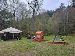 Uma tenda, um espaço de brincadeira infantil e um baloiço numa área gramada, com árvores ao fundo.