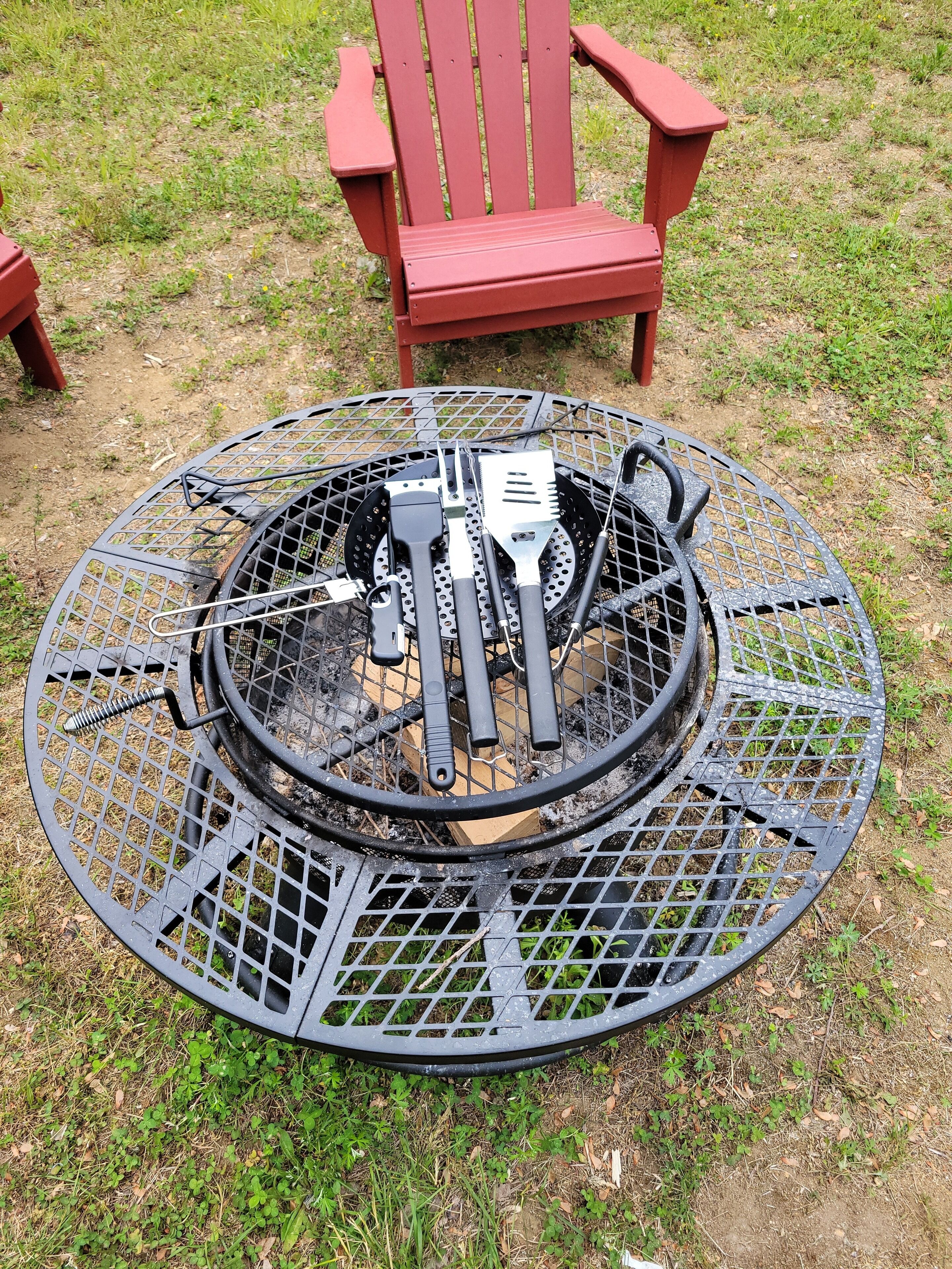 A round metal fire pit with grilling tools and a red chair in the background.