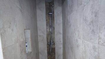A narrow shower area with a tiled floor, a wooden mat at the entrance, and a showerhead with a handheld shower attachment.