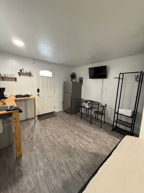 A compact room with a kitchenette, a dining area, a TV mounted on the wall, and a built-in shelf unit.