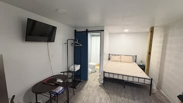 A small, compact room with a bed, a desk, a chair, a TV mounted on the wall, and a clothes drying rack.