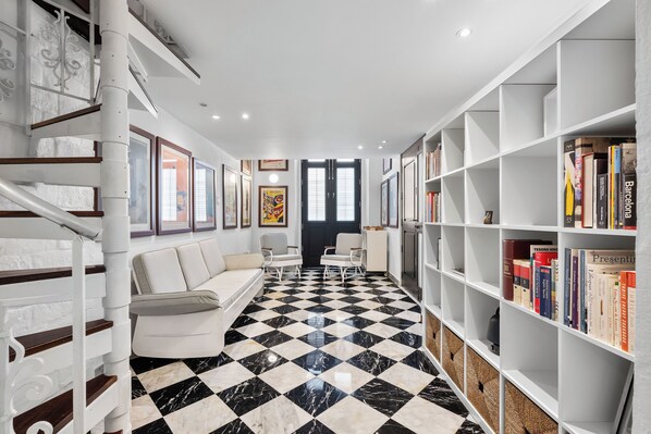 A hallway with a checkered floor, white walls, and a staircase on the left. There are two white armchairs and a bookshelf filled with books.