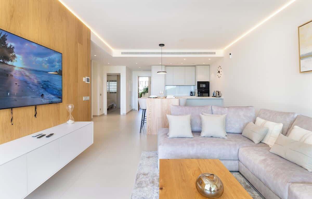 A modern living room with a large flat-screen TV, a sofa, a coffee table, and a kitchen area in the background.