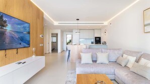 Ein modernes Wohnzimmer mit großem Flachbildfernseher, einer Couch, einem Couchtisch und einer Küchenzeile im Hintergrund.