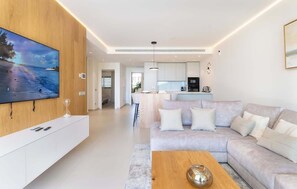 Een moderne woonkamer met een grote flatscreen-tv, een bank, een salontafel, en een keuken in de achtergrond.