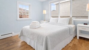 A bedroom with a bed, two bedside tables, a lamp, and a window with blinds. - Magnolia Portside – Spacious 3BR on Thames St (Newport)
