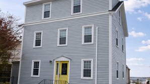 A two-story building with a yellow door, white window frames, and a brick foundation. - Magnolia Portside – Spacious 3BR on Thames St (Newport)