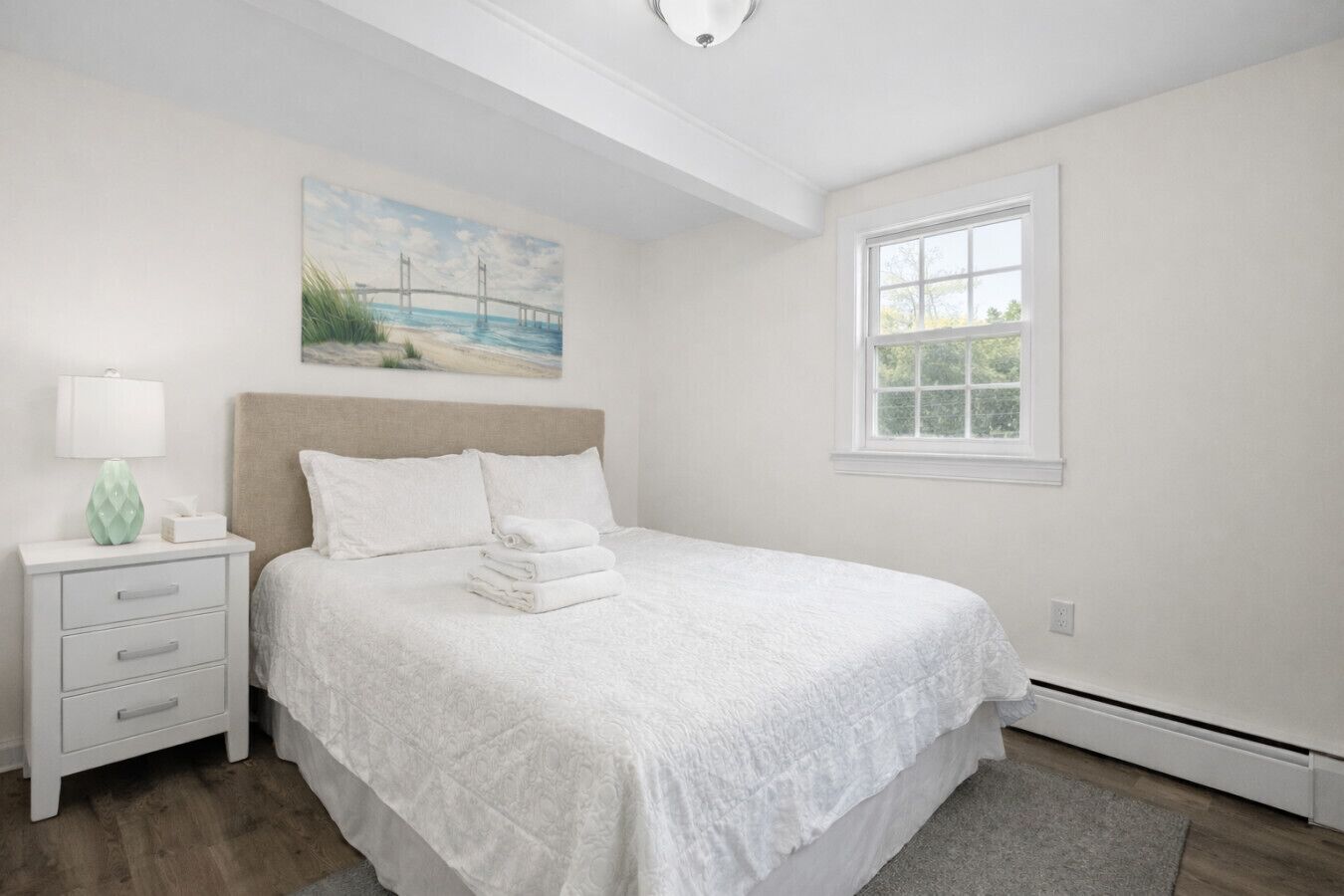 A bedroom with a bed, a nightstand, a lamp, a painting, and a window.