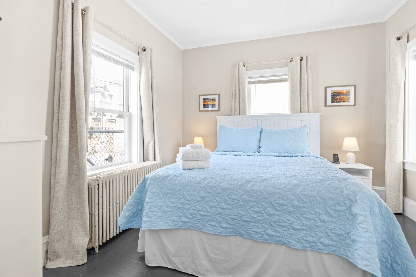Ein Schlafzimmer mit einem Bett, zwei Nachttischen, einem Fenster mit Vorhängen, einem Heizkörper und zwei gerahmten Bildern an der Wand.