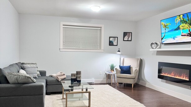 A modern living room with a grey sofa, a glass coffee table, a white rug, a fireplace, and a TV on the wall.