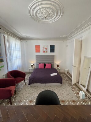 Une chambre d’hôtel avec un grand lit, deux tables de chevet, un fauteuil rouge, une chaise noire et un tapis à motifs.