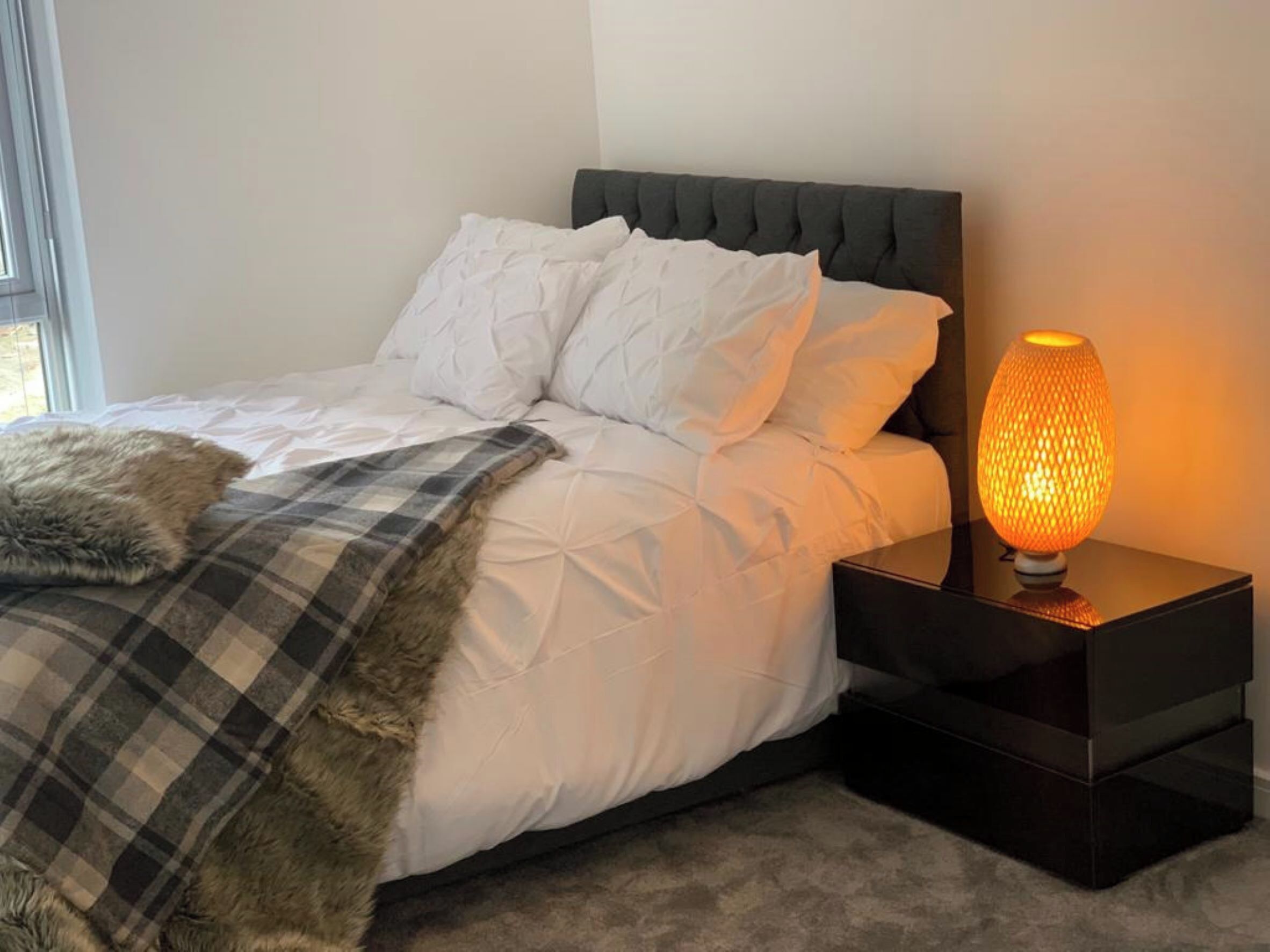 Ein ordentlich bezogenes Bett mit weißen Leinen und einem grauen Kopfteil, eine Karo-Decke und ein Fellüberwurf. Eine moderne Tischlampe mit strukturierter Lampenschirm steht auf einem schwarzen Nachttisch neben dem Bett.