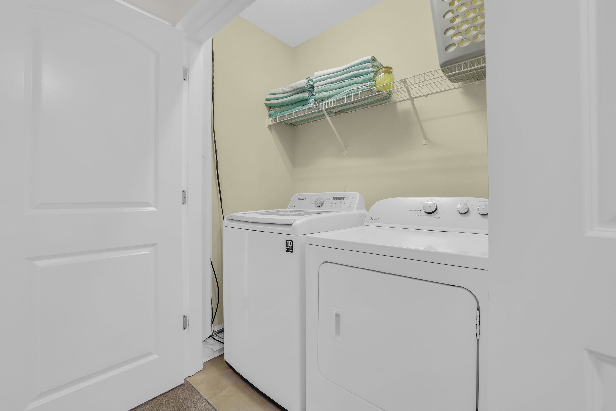 A laundry room with two white washers and dryers, a towel rack with folded towels, and a shelf with a glass container.