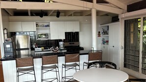A kitchen with white cabinets, a stainless steel refrigerator, a microwave, and a stove. There is a ceiling fan, a window with a view of greenery, and a flag hanging above the kitchen. - Shibui Cove (St. Thomas)