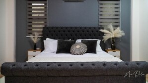 Quarto com cabeceira estofada, cama com almofadas brancas e estampadas, duas mesas de cabeceira com vasos decorativos e um aparelho de ar condicionado na parede.