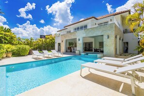 Un espace piscine avec des chaises longues, un bâtiment à deux étages et un ciel d’un bleu éclatant.