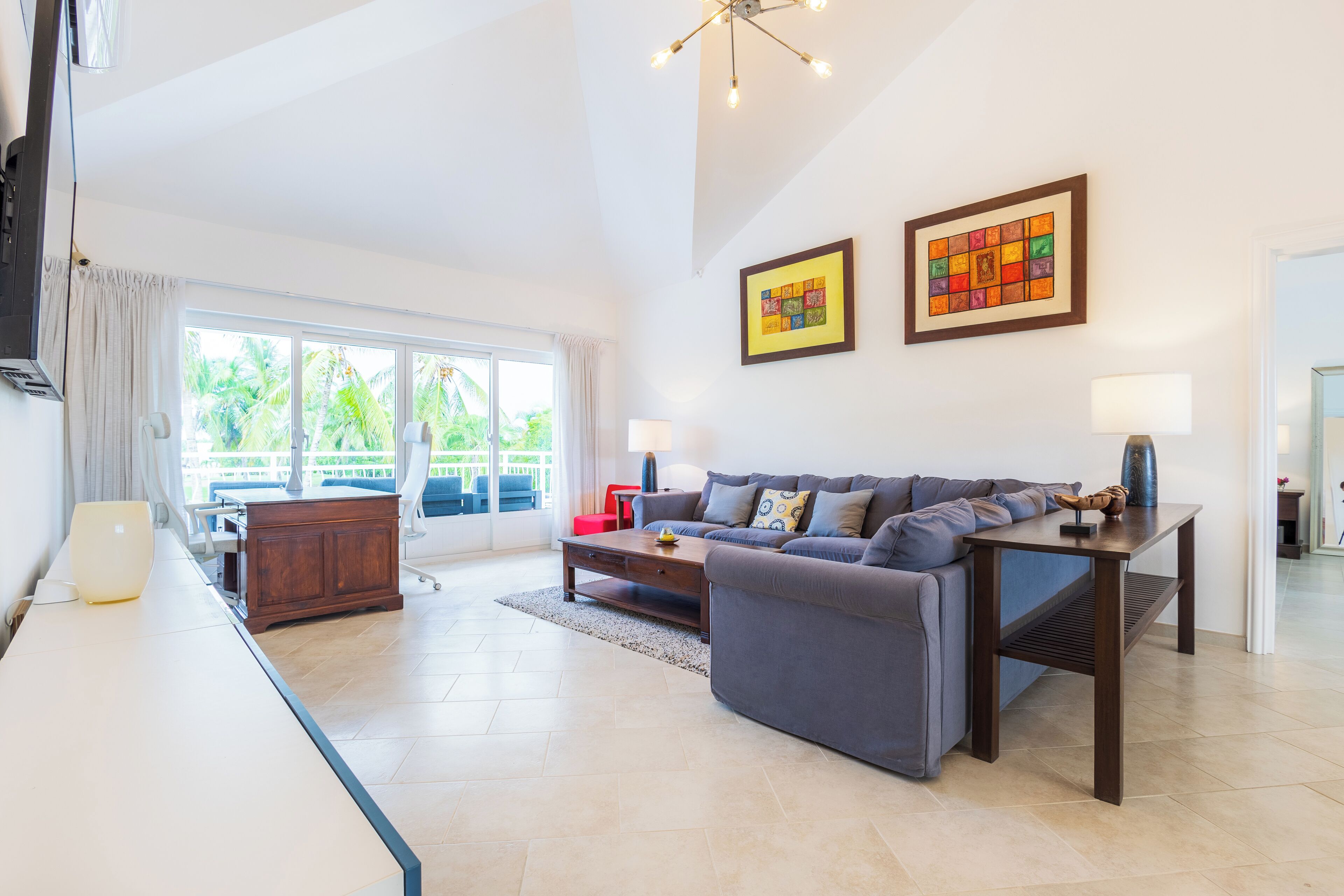 A spacious living room with a sofa, coffee table, and a TV. There are two framed artworks on the wall and a large window with a view of greenery.