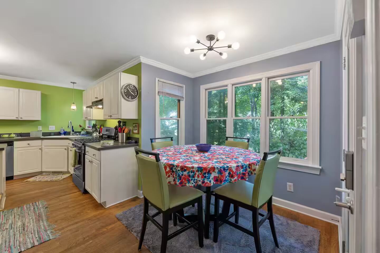 A kitchen with white cabinets, a dining table with a floral tablecloth, and a view of trees through large windows.