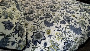 Une chambre à coucher avec un grand couvre-lit à motifs floraux, un ventilateur de plafond, une commode et une fenêtre avec des rideaux.