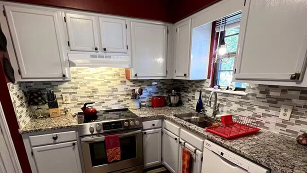 Une cuisine moderne avec des meubles blancs, un four en acier inoxydable, une bouilloire rouge et un lave-vaisselle.