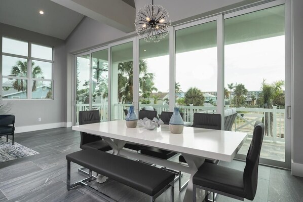 A spacious room with a large dining table, comfortable seating, and large windows offering a view of palm trees and a balcony.
