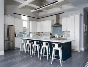 A modern kitchen with white cabinets, a large island with bar stools, a stainless steel refrigerator, and a blue-tiled backsplash.