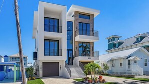 A modern multi-story residential building with a large garage, a balcony on the upper floor, and a small garden area with palm trees.
