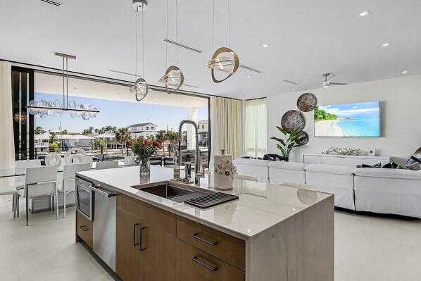 A modern kitchen with a central island, a dining area with chairs, a sofa, and a TV mounted on the wall.